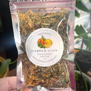 May include: A clear, sealed bag filled with dried herbs and flowers. The bag has a label that reads "GARDEN GLOW Simmer Pot Herb Scent Pack." The label features a floral and lemon design. The bag contains calendula, thyme, rosemary, lemon peel, and spearmint.