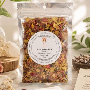 May include: A clear plastic pouch filled with dried flower petals and herbs labeled Aphrodites Aid Simmer Pot Herb Scent Pack 5 oz. The circular label lists ingredients including vanilla, rose, damiana, jasmine, calendula, brown sugar, nutmeg, hibiscus, and juniper berries.