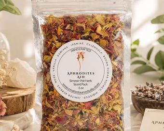 Puede incluir: Una bolsa de plástico transparente llena de pétalos de flores secas y hierbas etiquetada como Aphrodites Aid Simmer Pot Herb Scent Pack 142g. La etiqueta circular enumera ingredientes que incluyen vainilla, rosa, jazmín, caléndula, azúcar moreno e hibisco.