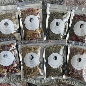 May include: Eight clear pouches filled with dried herbs and flowers, each labeled with a phase of the moon. The pouches are arranged on a decorative silver tray. The labels include text such as "New Moon" and "Waxing Crescent".