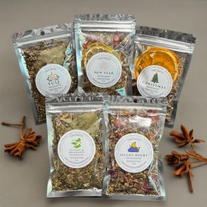 May include: Five clear pouches filled with dried herbs and botanicals. Each pouch has a circular label with text, including "Yule," "New Year," "Christmas," "Success & Abundance," and "Silent Night." Dried orange slices and star anise are also visible.
