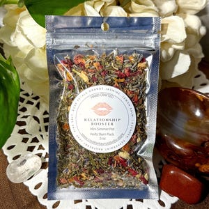 May include: A small clear pouch containing a blend of dried herbs and flowers labeled "Relationship Booster" with the text "Mini Simmer Pot Herb/Burn Pack 5 oz" and a website address.