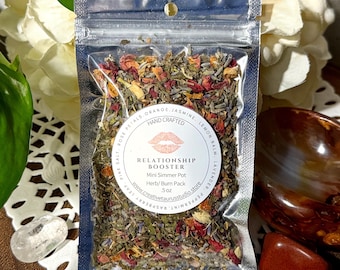 Puede incluir: Una pequeña bolsa transparente que contiene una mezcla de hierbas y flores secas etiquetada como "Relationship Booster" con el texto "Mini Simmer Pot Herb/Burn Pack 5 oz" y una dirección de sitio web.