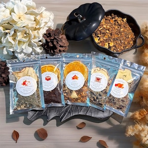 May include: An overhead shot showcases five clear pouches of potpourri with labels like "Apple Crumble" and "Coffee Cake." A black pumpkin-shaped container holds more potpourri. The scene is decorated with fall-themed elements, including a pinecone and dried leaves.