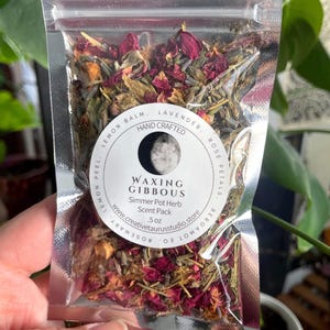 May include: A clear, sealed bag filled with dried herbs and rose petals. The bag has a circular label with the words "Waxing Gibbous" and "Simmer Pot Herb Scent Pack." The label also includes the website address www.creativetaurusstudio.store.