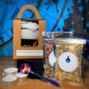 May include: A brown cardboard box with a clear window showing a white ceramic simmer pot and stand. The box is labeled "Mini Ceramic Simmer Pot & Stand with Tea Lights". Two small bags of dried herbs are in the foreground. One bag is labeled "DREAM WORK Mini Simmer Pot Herb Pack" and the other bag is labeled with a list of herbs including chamomile, catnip, hops, comfrey, and lavender. A rainbow iridescent spoon and four white tea lights are also in the image.