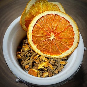 May include: A white ceramic bowl filled with a mixture of dried brown spice pieces and orange peel bits, topped with two large, circular, dehydrated orange slices that display a vibrant orange colour and a detailed, translucent citrus pulp texture.