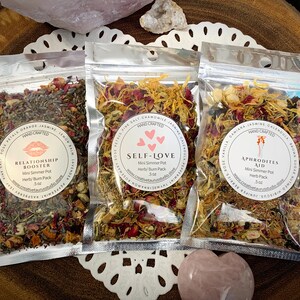 May include: Three small silver pouches containing dried herbs and flowers. Each pouch has a label with a different name and description: "Relationship Booster", "Self-Love", and "Aphrodite's Aid". Each pouch is labeled "Hand Crafted" and includes the size of the herb pack.