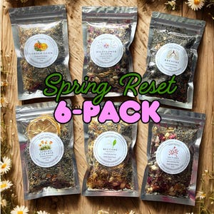 May include: Six sealed pouches of herbal blends, labeled with names like "Garden Glow" and "Wildflower Bloom." The text "Spring Reset 6-PACK" is displayed in green and pink. The pouches are arranged on a wooden surface, surrounded by daisies.