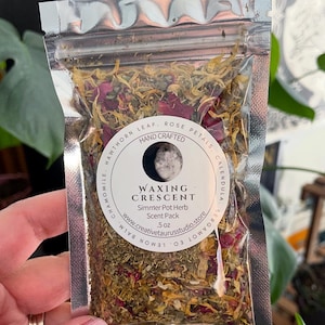 May include: A clear, sealed bag filled with dried herbs and flower petals. The bag has a label that reads "Waxing Crescent Simmer Pot Herb Scent Pack." The label also includes the words "Hand Crafted" and lists ingredients like chamomile and rose petals.
