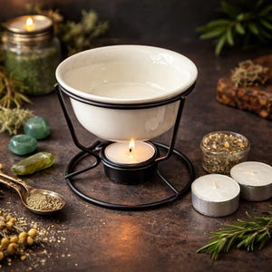 May include: A white ceramic essential oil diffuser bowl with a black metal stand, lit by a small candle. The setup includes small jars of herbs, green stones, and a spoon with herbs, creating a relaxing atmosphere.