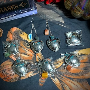 May include: Five silver heart-shaped tea infusers with different colored stones hanging from chains. The infusers are packaged in clear plastic bags.