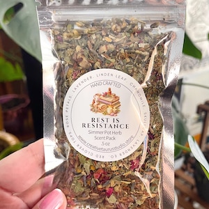 May include: A clear plastic bag filled with dried herbs and flowers. The bag has a circular label with the text "REST IS RESISTANCE" and "Simmer Pot Herb Scent Pack". The label also includes the website address www.creativetaurusstudio.store and the weight, .5 oz.