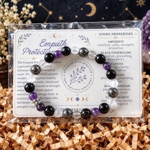 May include: A circular beaded bracelet featuring alternating polished spheres of purple amethyst, clear quartz, black tourmaline, and metallic hematite, presented on a rectangular card labeled Empath Protection with stone property text.