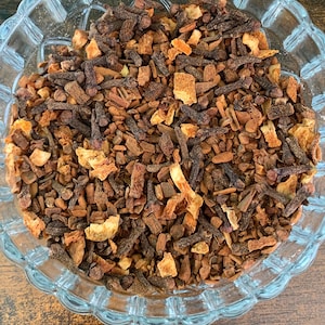 May include: A clear glass bowl filled with a mixture of dried brown cloves, small pieces of cinnamon bark, and dried orange peel. The ingredients are textured and varied in size, creating a rustic blend of dark brown and light orange tones.