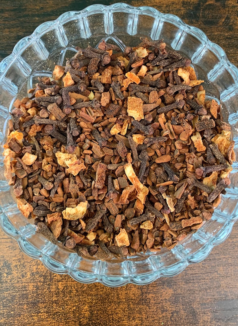 May include: A clear glass bowl filled with a mixture of dried brown cloves, small pieces of cinnamon bark, and dried orange peel. The ingredients are textured and varied in size, creating a rustic blend of dark brown and light orange tones.