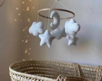 Op de afbeelding: Een baby mobile met witte pluche wolken, sterren en een maan, opgehangen aan een houten ring. De mobile hangt boven een gevlochten mand. De achtergrond is een zachte, neutrale kleur.