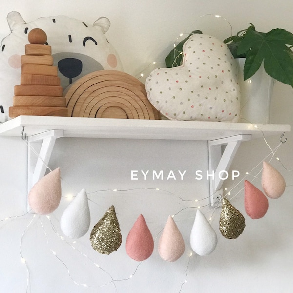 Può includere: Una ghirlanda di gocce di pioggia in feltro rosa, bianco e oro con una stringa di luci. La ghirlanda è appesa a uno scaffale bianco con un arcobaleno di legno, un peluche e un cuscino a forma di cuore sullo scaffale. Il testo "EYMAY SHOP" è visibile sulla ghirlanda.
