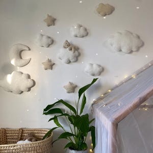 May include: A nursery wall decoration featuring felt clouds, stars, and a crescent moon. A small teddy bear rests on one cloud. A woven basket and a potted plant are in the foreground. The scene is illuminated by fairy lights.