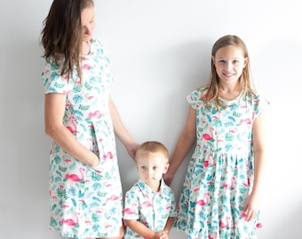 May include: A family wearing matching white dresses and shirts with a pink flamingo and tropical leaf pattern. The dresses and shirts have short sleeves. The woman is wearing a white dress with a pink flamingo and tropical leaf pattern. The girl is wearing a white dress with a pink flamingo and tropical leaf pattern. The boy is wearing a white shirt with a pink flamingo and tropical leaf pattern.