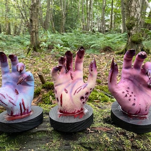 May include: Three realistic-looking severed hands with long, sharp nails, painted in shades of blue, pink, and white, with red blood dripping from them. The hands are mounted on black wooden bases.