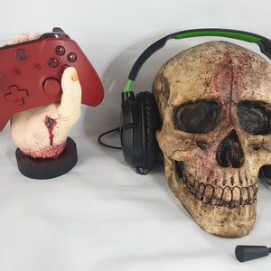 May include: A red video game controller in a bloody hand-shaped stand next to a black and green gaming headset on a skull.