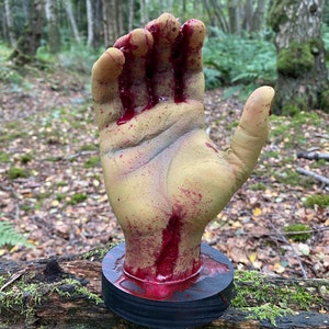 May include: A gruesome Halloween prop featuring a severed hand with a yellow-green skin tone, splattered with fake blood. The hand is mounted on a black circular base, creating a spooky and realistic effect for Halloween decorations.
