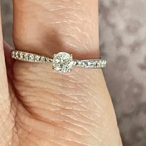 K&ouml;nnte beinhalten: Ein silberner Verlobungsring mit einem einzelnen runden Diamanten und einer Reihe kleinerer Diamanten.