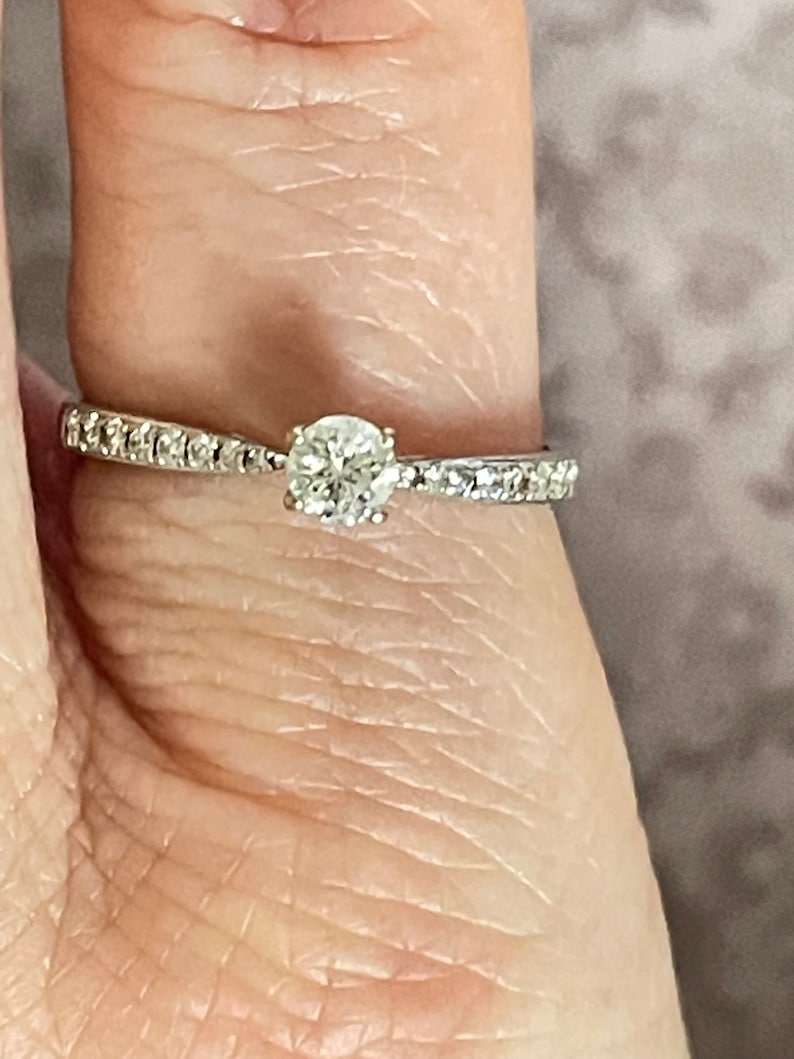 K&ouml;nnte beinhalten: Ein silberner Verlobungsring mit einem einzelnen runden Diamanten und einer Reihe kleinerer Diamanten.