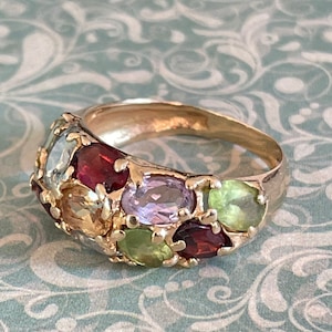 Könnte beinhalten: Ein goldener Ring mit einer Reihe von bunten Edelsteinen. Die Steine sind in einer Reihe um die Band herum gesetzt und umfassen grüne, rote, violette und blaue Steine.