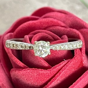 K&ouml;nnte beinhalten: Ein silberner Ring mit einem einzigen runden Diamanten, der in der Mitte sitzt. Der Ring ist von kleineren Diamanten umgeben und liegt auf einem roten Samt-Hintergrund.