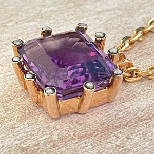 May include: A gold pendant necklace with a large purple amethyst gemstone set in a gold frame with small pearl accents.