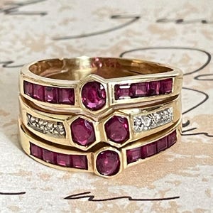 May include: Three gold rings stacked together, featuring a combination of round and rectangular red gemstones. One ring has small, clear stones. The rings are set against a patterned background.