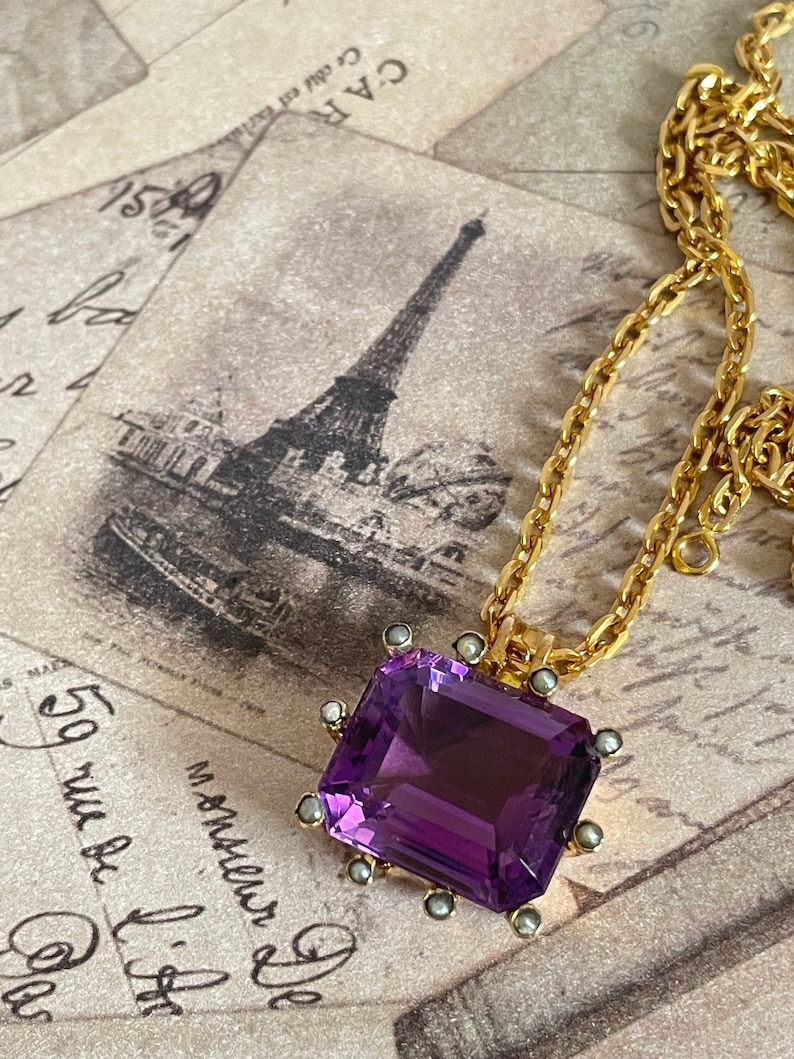 May include: A gold chain necklace with a large, rectangular, faceted amethyst pendant. The pendant is set in a gold frame with small pearl accents.