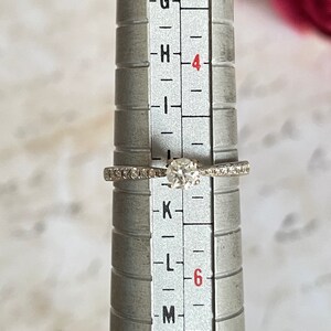K&ouml;nnte beinhalten: Ein silberner Ring mit einem runden, klaren Diamanten und kleinen Diamanten, die in einer Bandfassung sitzen. Der Ring ist auf einem Ringma&szlig;stab kalibriert und zeigt eine Gr&ouml;&szlig;e von 6.