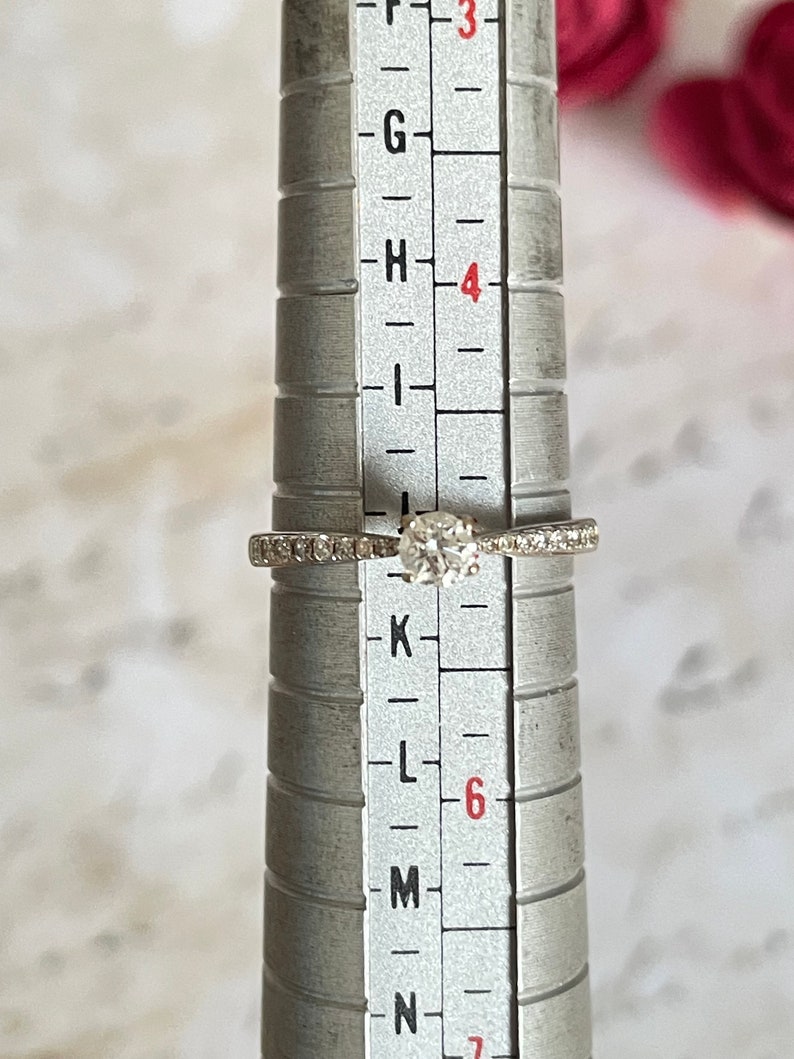 K&ouml;nnte beinhalten: Ein silberner Ring mit einem runden, klaren Diamanten und kleinen Diamanten, die in einer Bandfassung sitzen. Der Ring ist auf einem Ringma&szlig;stab kalibriert und zeigt eine Gr&ouml;&szlig;e von 6.