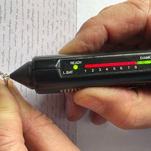 K&ouml;nnte beinhalten: Ein schwarzer Diamantentester mit einem gr&uuml;nen Licht, das anzeigt, dass ein Diamant vorhanden ist. Der Tester hat eine rote Leiste mit den Zahlen 1 bis 8 und die W&ouml;rter "READY" und "L.BAT" darauf.