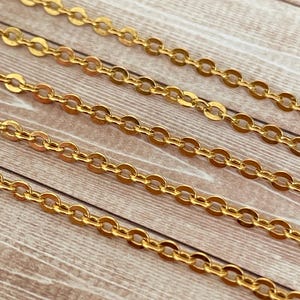 May include: Four strands of gold chain on a wooden surface. The chain is made of small, oval-shaped links.