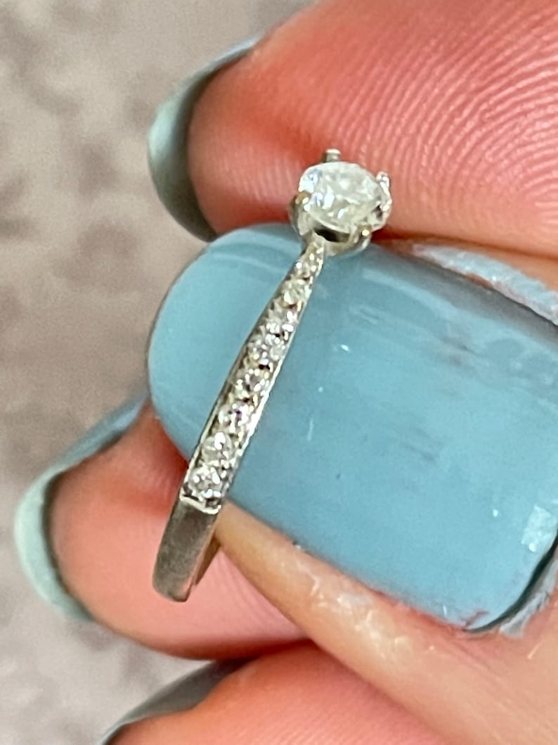K&ouml;nnte beinhalten: Ein silberner Ring mit einem einzelnen runden Diamanten in der Mitte und kleineren Diamanten, die in einer Band um den Ring gesetzt sind.