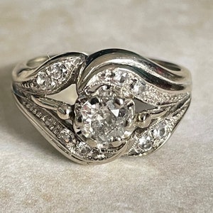 Könnte beinhalten: Ein silberner Ring mit einem großen, runden, klaren Diamanten in der Mitte. Der Ring ist mit kleineren Diamanten besetzt, die den Mitteldiamanten umgeben und entlang des Rings verlaufen. Der Ring hat ein zartes, filigranes Design.