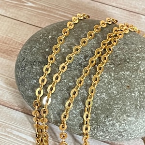 May include: A close-up of a gold chain necklace with a delicate, oval-shaped link pattern. The chain is draped over a gray stone, showcasing its intricate design.