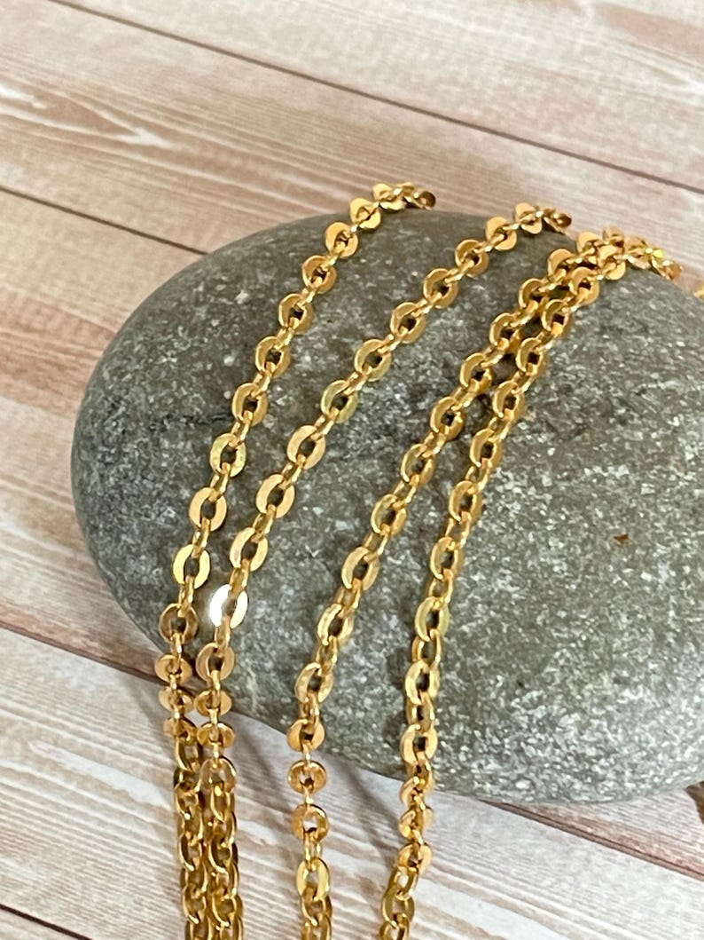 May include: A close-up of a gold chain necklace with a delicate, oval-shaped link pattern. The chain is draped over a gray stone, showcasing its intricate design.