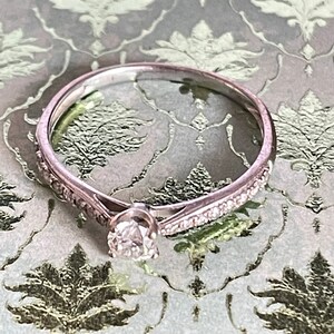 K&ouml;nnte beinhalten: Ein zarter Silberring mit einem einzelnen runden Diamanten in der Mitte und einer Reihe kleinerer Diamanten entlang des Bandes.