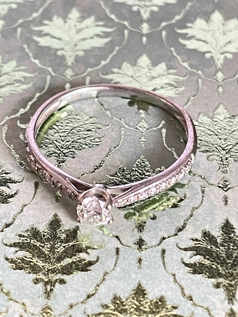 K&ouml;nnte beinhalten: Ein zarter Silberring mit einem einzelnen runden Diamanten in der Mitte und einer Reihe kleinerer Diamanten entlang des Bandes.