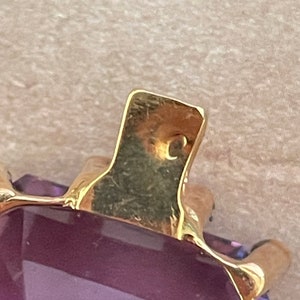 May include: Close-up of a gold pendant with a large, faceted, purple amethyst gemstone. The pendant has a simple, rectangular shape and a smooth, polished finish.