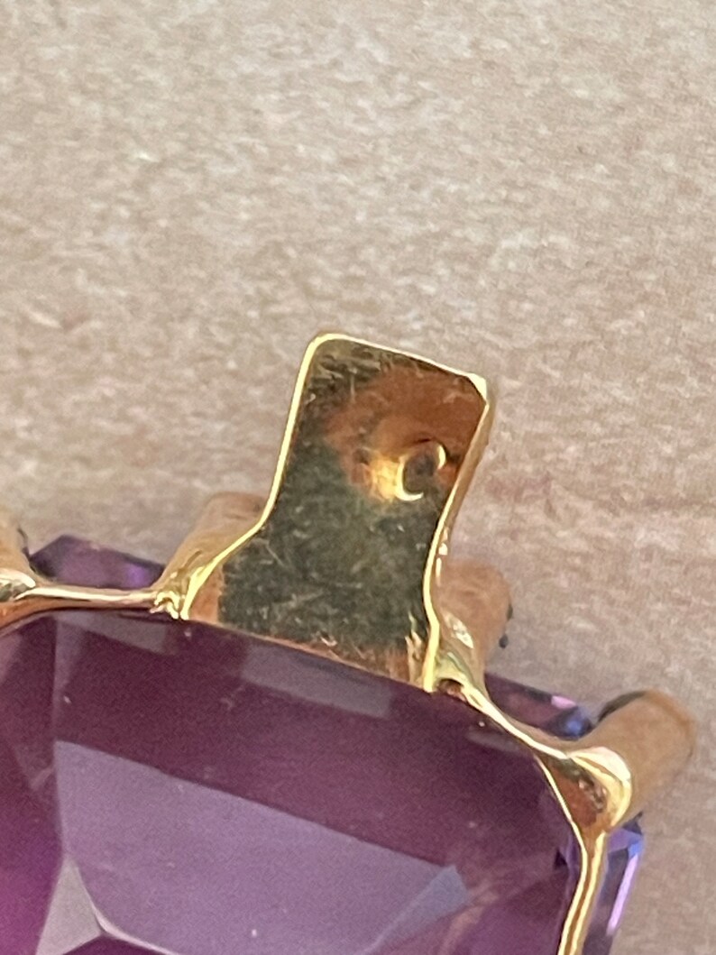 May include: Close-up of a gold pendant with a large, faceted, purple amethyst gemstone. The pendant has a simple, rectangular shape and a smooth, polished finish.