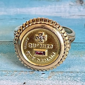 Könnte beinhalten: Ein goldfarbener Ring mit einem kreisförmigen Gesicht mit der Inschrift "QUE HIER QUE DEMAIN". Der Ring hat ein Kreuzdesign mit einem kleinen Diamanten und einem rechteckigen Rubin. Das Band des Rings ist silberfarben.