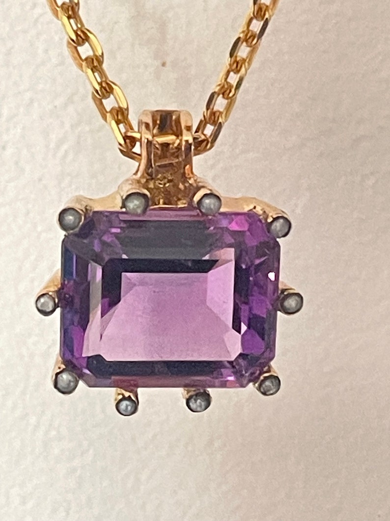 May include: A gold chain necklace with a pendant featuring a large, square-shaped amethyst gemstone surrounded by small pearl accents.