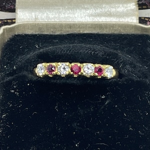 May include: A gold band ring with alternating red and white gemstones. The ring is in a black velvet ring box.