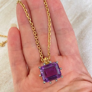 May include: A gold chain necklace with a large, rectangular, purple amethyst pendant. The pendant is set in a gold frame with small, white pearls around the edges.